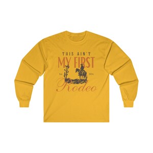 May include: Mustard yellow long-sleeve shirt with the text "THIS AIN'T MY FIRST Rodeo" in a western-style font. The design includes a silhouette of a cowboy on a horse, a cactus, and the year 2024.
