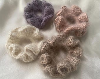 May include: A collection of four circular hair scrunchies made from fuzzy, textured yarn. The set includes one purple, one light pink, one cream, and one beige scrunchie, each featuring a ruffled, gathered design with a soft, fibrous appearance.