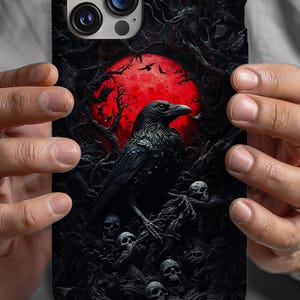 May include: A black phone case with a dark forest scene featuring a crow, a red moon, and skulls. The case has a camera lens cutout.