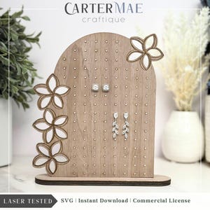 Puede incluir: Soporte de exhibición de aretes de madera con un diseño floral. El soporte tiene una parte superior de arco redondeado y una base. Tiene múltiples agujeros para sujetar aretes. El soporte está cortado con láser y tiene el texto "CARTERMAE" y "craftique" en él.