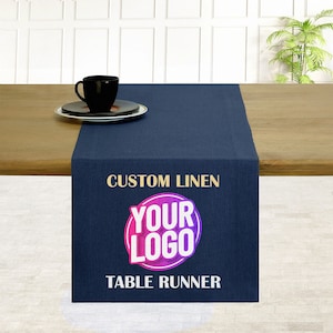 May include: A navy blue linen table runner with gold text that reads "Custom Linen Table Runner". A circle with the text "Your Logo" in a pink and purple gradient is centred on the runner.