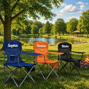 May include: Three personalized camping chairs in blue, orange, and black are set up on a grassy lawn. Each chair has a name printed on the backrest: "Sophie", "Thomas", and "Blake". The chairs have silver frames and armrests.