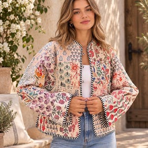 May include: A long-sleeved quilted jacket featuring a patchwork design with various floral and botanical patterns in shades of pink, red, blue, and green. The jacket has a round neckline, a black and white striped trim, and two front pockets.
