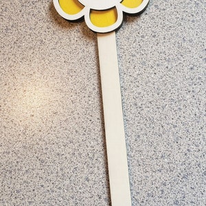 May include: A wooden flower stake with a yellow painted flower and a smiling face. The flower has a white center and blue eyes.