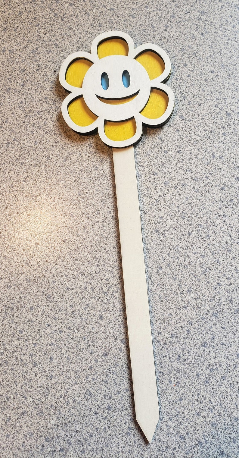 May include: A wooden flower stake with a yellow painted flower and a smiling face. The flower has a white center and blue eyes.