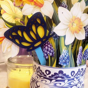 May include: A blue and yellow butterfly cutout with a blue stick attached to it. The butterfly is in a blue and white ceramic vase with a floral pattern. The vase is filled with a bouquet of yellow and white daffodils and purple grape hyacinths.