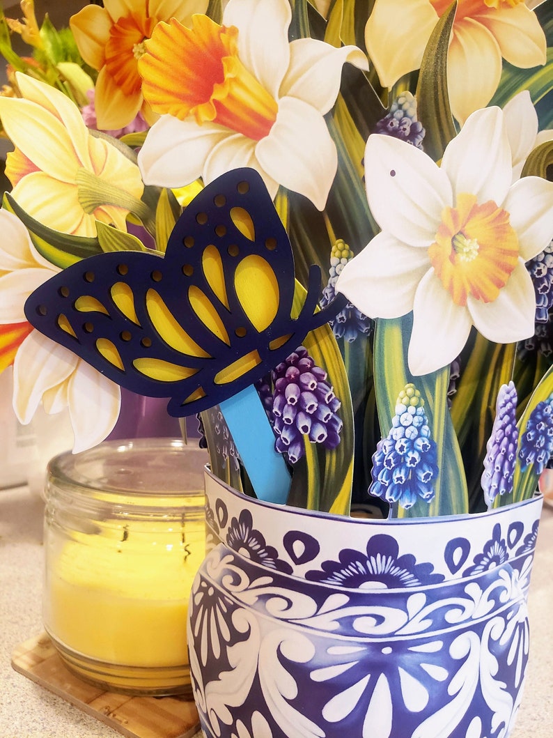 May include: A blue and yellow butterfly cutout with a blue stick attached to it. The butterfly is in a blue and white ceramic vase with a floral pattern. The vase is filled with a bouquet of yellow and white daffodils and purple grape hyacinths.