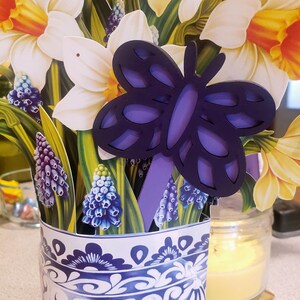 May include: A blue and white ceramic vase with a floral pattern, filled with a bouquet of yellow daffodils and blue grape hyacinths. A purple butterfly cutout is placed in the vase.