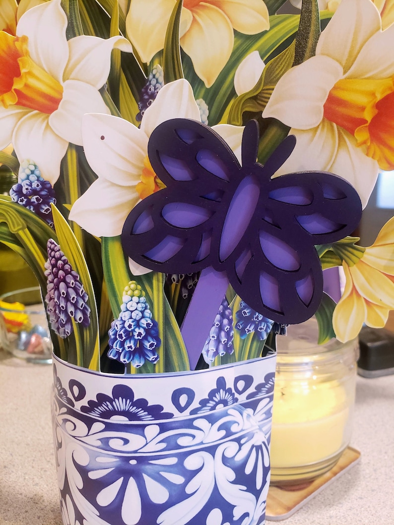 May include: A blue and white ceramic vase with a floral pattern, filled with a bouquet of yellow daffodils and blue grape hyacinths. A purple butterfly cutout is placed in the vase.