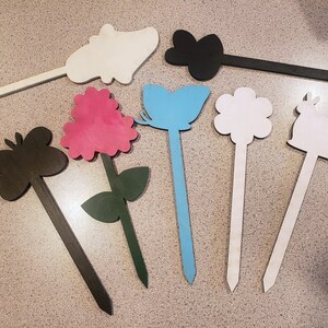 May include: A set of eight wooden garden stakes with various shapes and colors, including a butterfly, flower, rabbit, and a pink flower. The stakes are painted in black, white, blue, and pink.