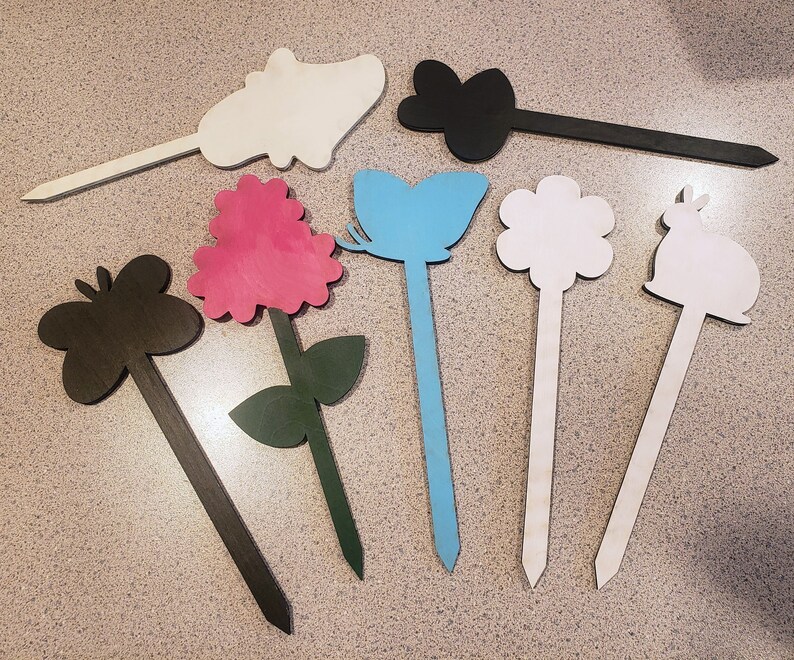 May include: A set of eight wooden garden stakes with various shapes and colors, including a butterfly, flower, rabbit, and a pink flower. The stakes are painted in black, white, blue, and pink.