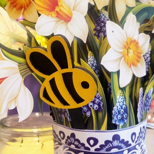 May include: A white ceramic vase with a blue and white floral pattern, filled with yellow and white daffodils and purple grape hyacinths. A wooden bee cutout is placed in the vase.