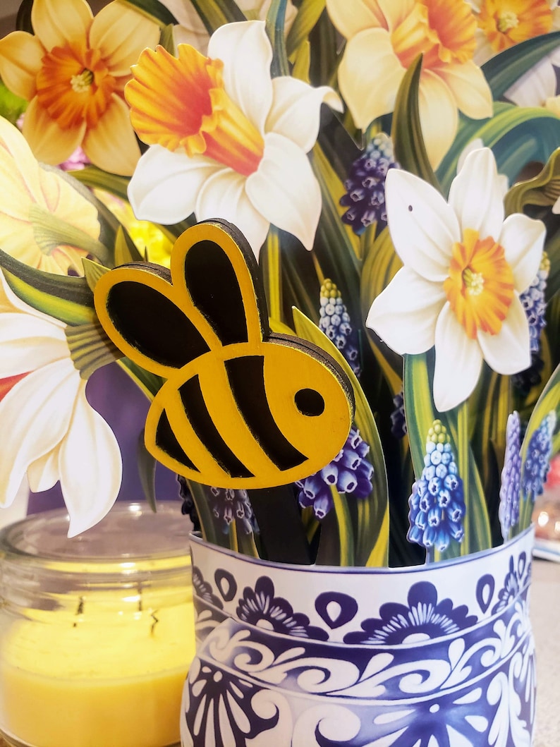 May include: A white ceramic vase with a blue and white floral pattern, filled with yellow and white daffodils and purple grape hyacinths. A wooden bee cutout is placed in the vase.