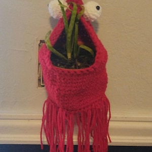 Może przedstawiać: Czerwony, dziany wiszący pojemnik na rośliny z frędzlami, z wypukłymi oczami i małą zieloną rośliną w środku.