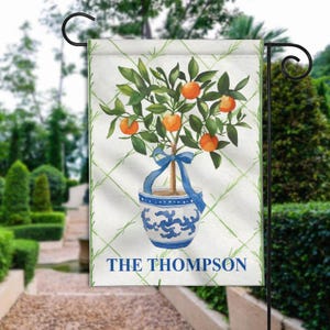 May include: A white garden flag with a green and white checkered pattern. A watercolor illustration of an orange tree in a blue and white planter with a blue ribbon. The text "THE THOMPSON" is printed at the bottom of the flag.