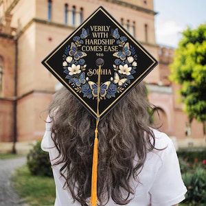 May include: A black graduation cap with gold trim and a tassel. The cap features a floral and butterfly design in blue and white, with the text "VERILY WITH HARDSHIP COMES EASE" and the name "SOPHIA".