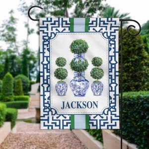 May include: A blue and white garden flag with a geometric pattern and a design of three topiary trees in blue and white ceramic pots. The flag has the name "JACKSON" printed on it.