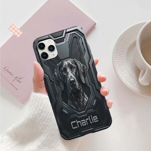 May include: A black smartphone case with a rugged, mechanical-style texture featuring a detailed illustration of a dark-colored dog's face in the center and the name Charlie printed in a bold, light-colored font at the bottom of the case.