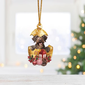 May include: A gold Christmas ornament with a brown and black dog peeking out of the top. The ornament is decorated with red and green holly, a red lantern, and a candy cane.