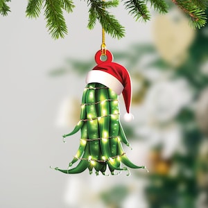 May include: A green string bean Christmas ornament with a red Santa hat and white trim. The string beans are wrapped with white lights.