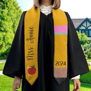 May include: A yellow graduation sash with a pencil design and glitter accents. The sash has the text "2024" and "Miss Annie" printed on it.