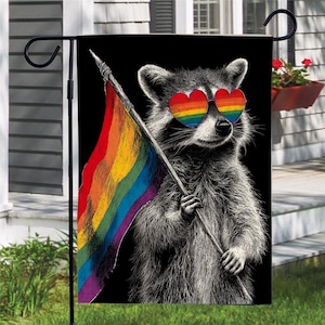 May include: A garden flag featuring a raccoon wearing heart-shaped rainbow sunglasses and holding a rainbow flag. The raccoon is depicted in black and white against a black background. The flag is displayed on a black metal pole.