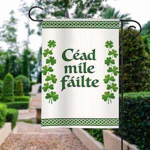 May include: A white garden flag with green Celtic knotwork and shamrock accents. The flag displays the words "Céad míle fáilte" in green, a traditional Irish greeting. The flag is hanging on a black metal garden stake.