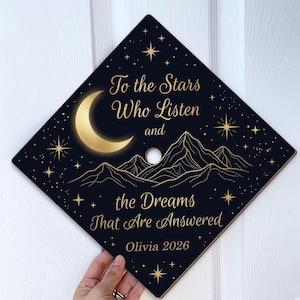 May include: A black graduation cap with gold celestial design. The cap features a crescent moon, stars, and mountains. Text reads "To the Stars Who Listen and the Dreams That Are Answered" and "Olivia 2026".