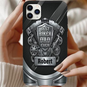 May include: A black phone case featuring a metallic engine graphic with a central shield displaying the text BEST BIKER DAD EVER and a rectangular nameplate below with the name Robert in bold black letters against a silver background.