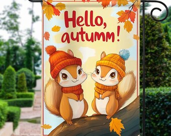 May include: A garden flag with a yellow background featuring two cartoon squirrels wearing hats and scarves. The squirrels are sitting on a tree branch with autumn leaves. The text "Hello, autumn!" is written in red and black letters.