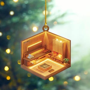 May include: A wooden ornament shaped like a sauna with a small fireplace, a bench, and towels. The ornament has a gold string for hanging and the word "NAME" is written on the side.