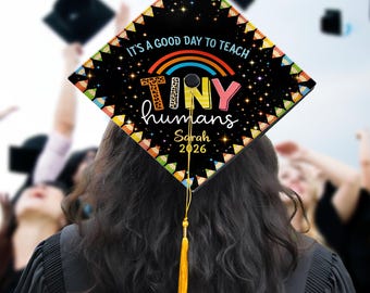 May include: A black graduation cap with the text "IT'S A GOOD DAY TO TEACH" and "TINY humans" in colorful letters. A rainbow and stars decorate the cap, with the name "Sarah" and the year "2026" below. A yellow tassel hangs from the cap.
