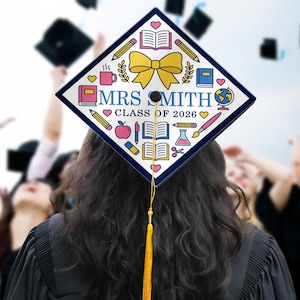 May include: A graduation cap with a white surface and a navy blue border. The cap is decorated with school-themed icons, a yellow bow, and the text "MRS SMITH CLASS OF 2026". A yellow tassel hangs from the cap.