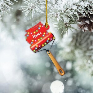 May include: Red paint roller ornament with a string of colorful lights and the word "Name" on it. The ornament is hanging from a gold string on a snowy pine branch.