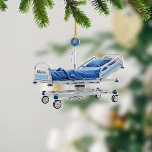 May include: A silver hospital bed ornament with blue sheets and a blue pillow. The bed has wheels and a name tag that says "Name".
