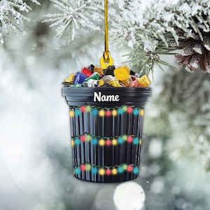 May include: A black trash can Christmas ornament with colorful lights and a name tag. The trash can is filled with colorful cans and bottles.