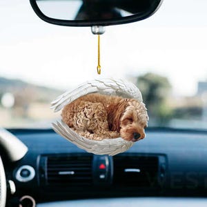May include: A white ceramic car charm with a small, sleeping brown and white dog inside a pair of angel wings. The charm is hanging from a rearview mirror in a car.