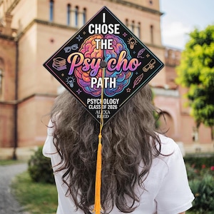 May include: A graduation cap with the words "I Chose The Psycho Path" in colorful lettering. The cap also includes the text "Psychology Class of 2026 Alexa Reed" and is decorated with various icons and a gold tassel.