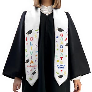 May include: A white graduation sash with colorful text and graphics. The sash features the name "Olivia" and the word "Kindergarten" on one side, and "GRADUATE" and "SCHOOL NAME" on the other. Graduation caps, stars, and confetti are also included.
