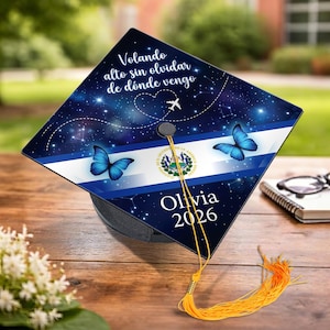 May include: A graduation cap with a dark blue background featuring a starry sky design, butterflies, and the text "Volando alto sin olvidar de dónde vengo." The cap includes the name "Olivia" and the year "2026."
