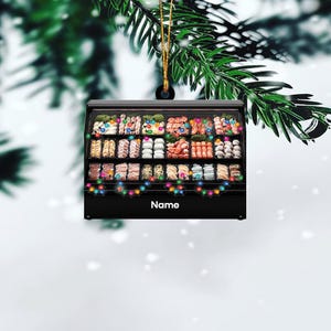 May include: A black Christmas ornament shaped like a refrigerated display case filled with various types of meat and seafood. The case is decorated with colorful string lights and the word "Name" is printed on the front.