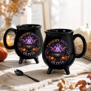 May include: Two black ceramic mugs shaped like cauldrons with three small feet and a handle. Each mug features a circular graphic with a bubbling cauldron, witch hats, pumpkins, and the text "WITCH'S BREW" and "OLIVIA" printed in purple and orange.