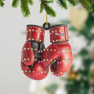 May include: Red boxing gloves Christmas ornament with string lights. The gloves have black accents and the word "Name" is printed on one of the gloves. The ornament is hanging from a gold string.