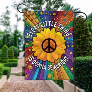 May include: A colorful garden flag with a rainbow background and a yellow sunflower with a peace sign in the center. The text "Every little thing is gonna be alright" is written in a circle around the sunflower.