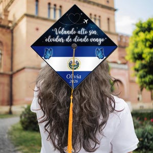 May include: A graduation cap featuring the flag of El Salvador, adorned with blue butterflies and the text "Volando alto sin olvidar de dónde vengo." The cap also includes the name "Olivia" and the year "2026."