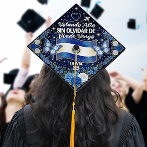 May include: A graduation cap with a navy blue background, adorned with blue butterflies, flowers, and the flag of El Salvador. The cap features the text "Volando Alto SIN OLVIDAR DE Dónde Vengo" and the name "Olivia 2026". A gold tassel hangs from the cap.