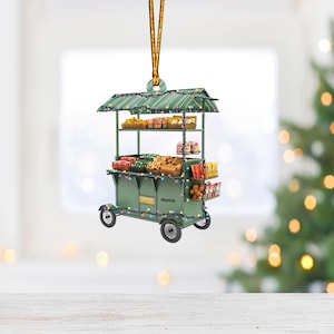May include: A green Christmas ornament shaped like a food cart with various snacks and sweets. The cart has a striped awning and is decorated with colorful string lights.  The cart has wheels and a sign that says 