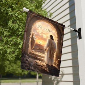 May include: A decorative outdoor flag featuring an artistic depiction of Jesus standing outside a tomb. The image includes the text "He is Risen" and a sunset backdrop with three crosses. The flag is mounted on a white pole.