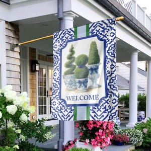 May include: A rectangular outdoor flag with a blue and white geometric pattern border. The center features a garden scene with potted topiary and the word "WELCOME". The flag is displayed on a porch with white columns and flowering plants.