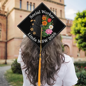 May include: Black graduation cap with the words "Social Worker" and "Linda MSW 2026". The cap features a brain graphic on one side and colorful flowers on the other. A gold tassel hangs from the cap.